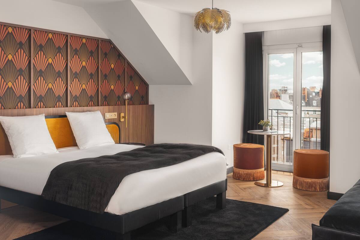 A modern bedroom with a bed, a wooden headboard, a hanging light fixture, and a view of the city through a large window.