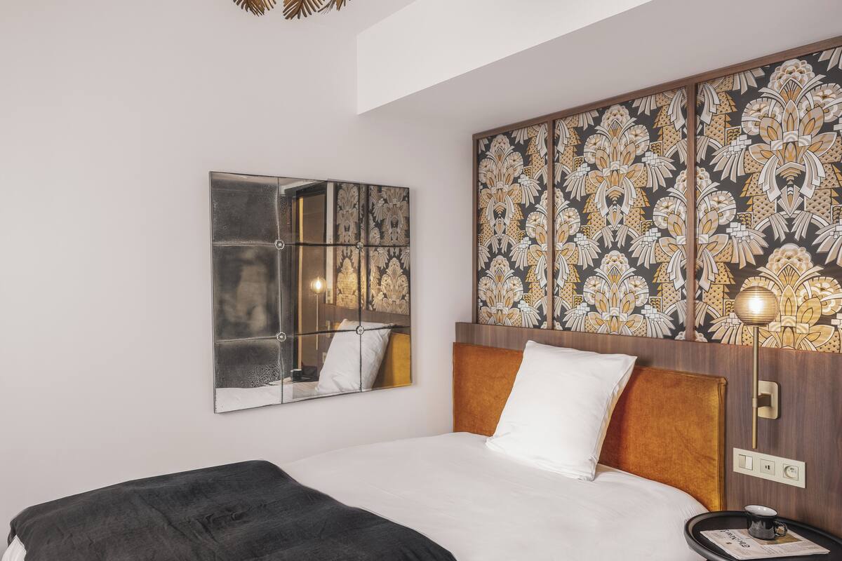 A hotel room with a bed, a bedside table with a lamp, a wall-mounted mirror, and a decorative panel with a floral pattern.