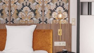 A hotel room with a bed, a round table with a lamp, a patterned wallpaper, and a door.