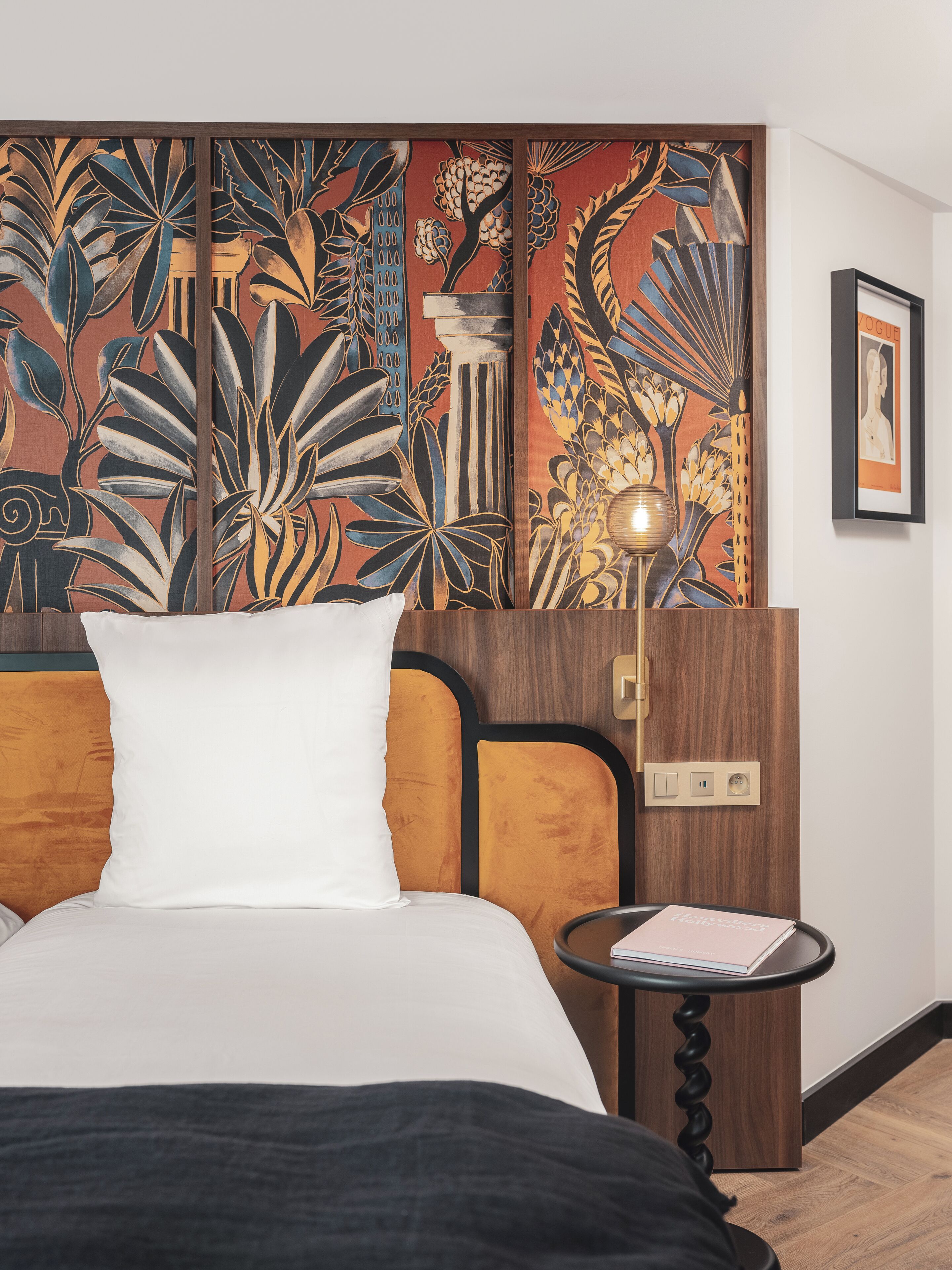 Habitación de hotel con cama, mesita de noche, lámpara y un mural floral en la pared.
