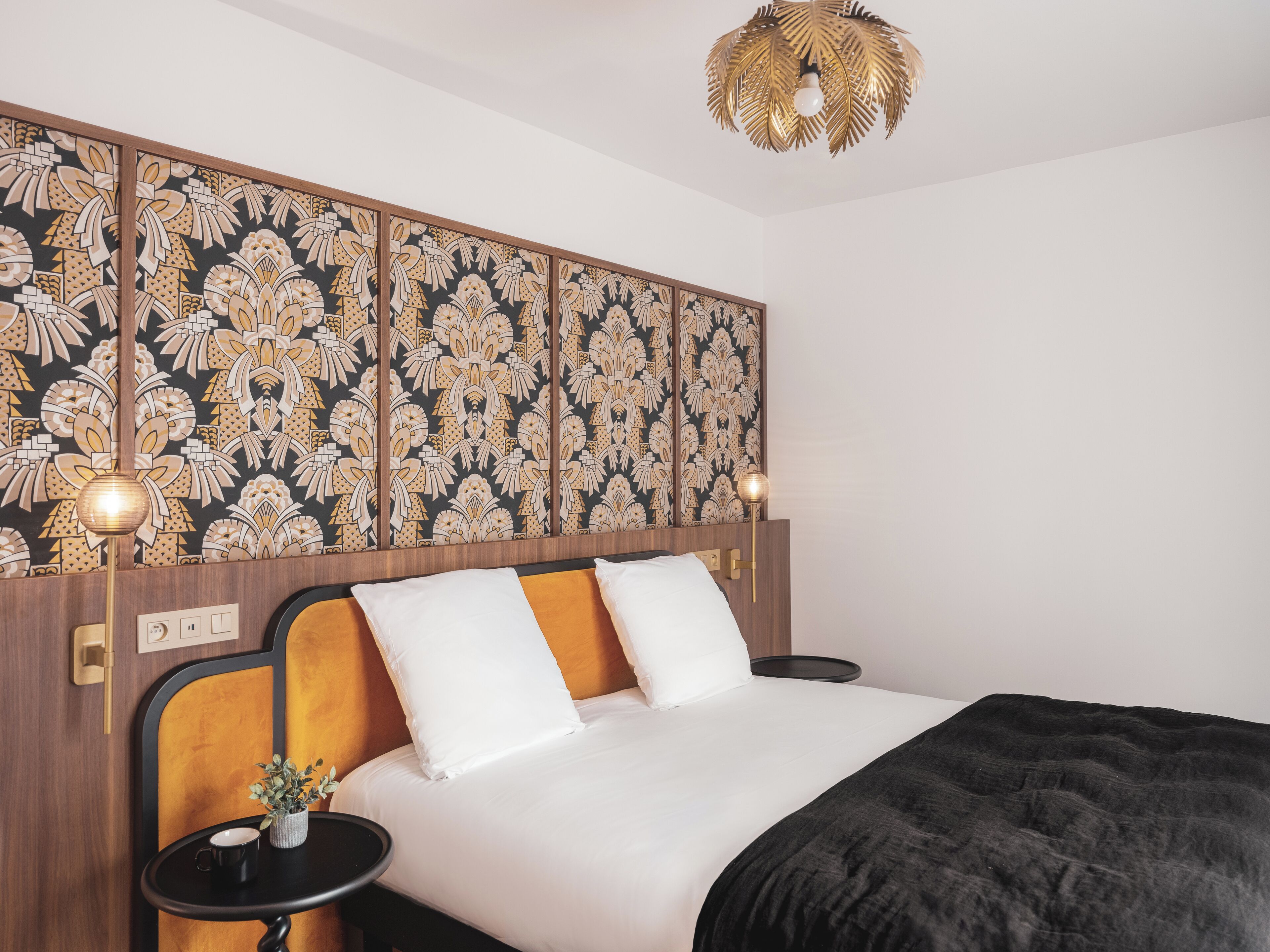 A hotel room with a bed, bedside tables, a decorative headboard, a wall with a floral pattern, and a ceiling light fixture.