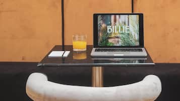 Een laptop op een tafel met een glas sinaasappelsap, een notitieboekje en een pen. Op de achtergrond is een gestreepte muur en een zachte bank te zien.