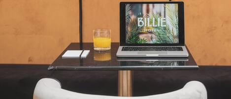 A laptop on a table with a glass of orange juice, a notepad, and a pen. The background features a striped wall and a cushioned bench.