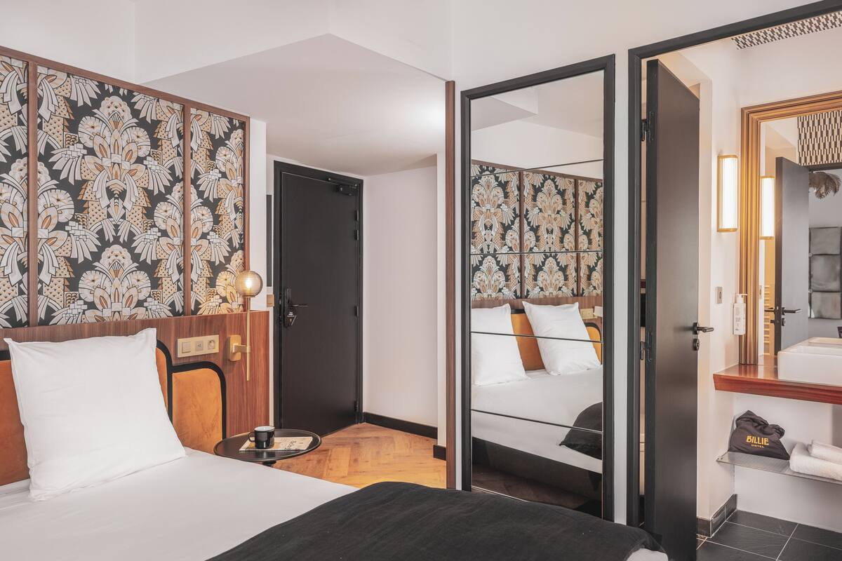 A hotel room with a bed, a bedside table with a cup, a mirror, a bathroom with a sink and towel rack.