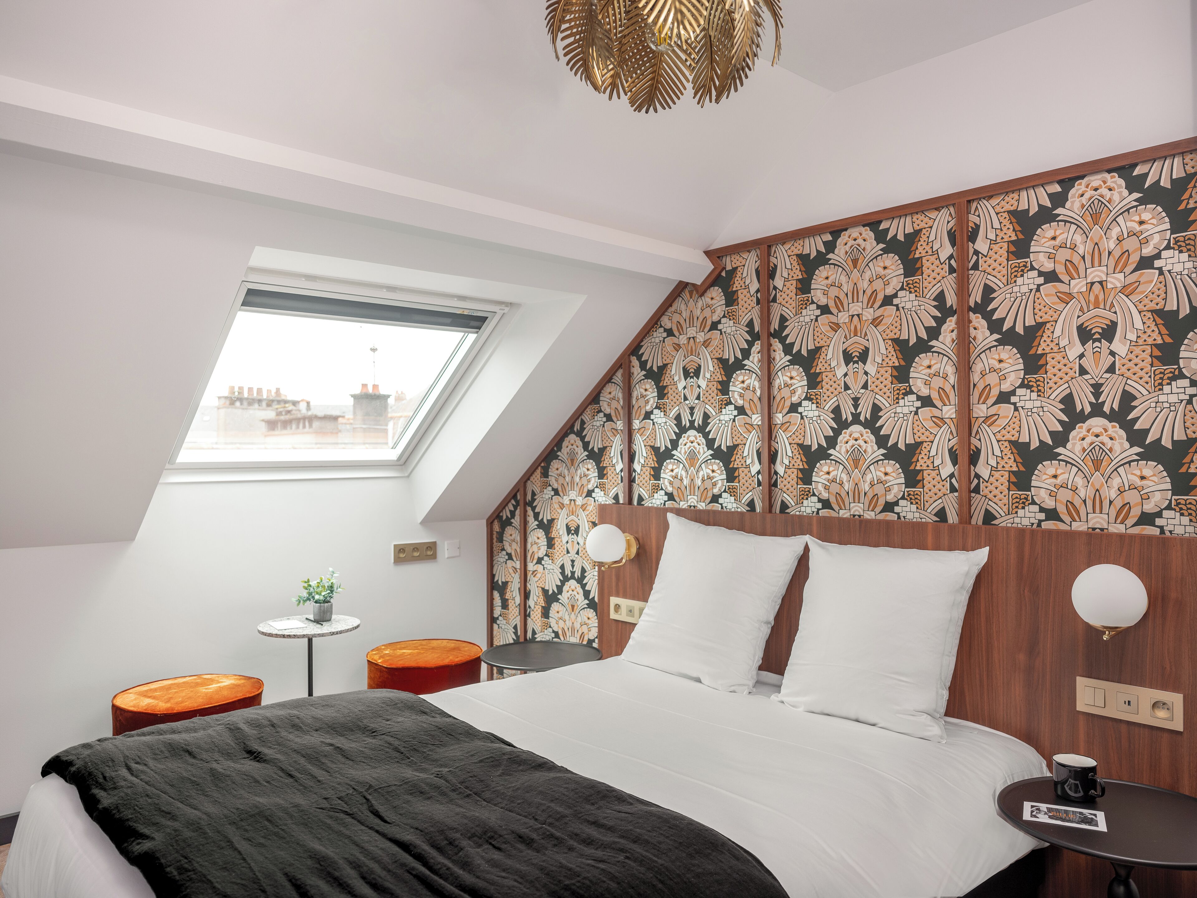 A modern bedroom with a sloped ceiling, a skylight, a bed with white bedding, a small round table with a cup, and a decorative wall panel.