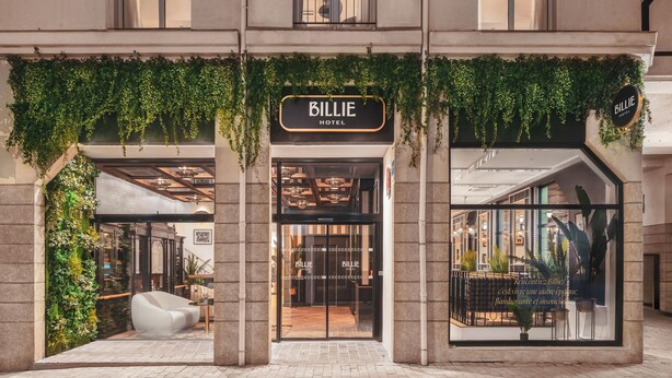 L’entrée d’un hôtel, avec une enseigne indiquant « Billie Hotel », présente un intérieur moderne avec une façade en verre et des plantes d’intérieur.