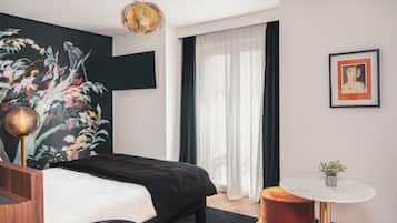 A modern bedroom with a bed, a round side table, a small round table, a hanging light fixture, a framed picture, and a large floral mural on the wall.