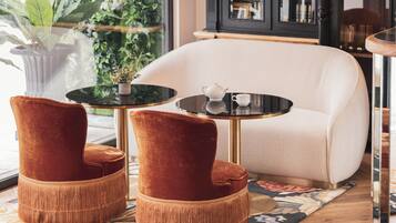 Una sala de estar moderna con un sillón blanco, dos sillones tapizados en terciopelo rojo, una mesita redonda con una tetera y una taza blancas, y un ventanal grande con follaje en el exterior.