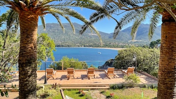 Terraço com bancos de madeira, vista para a costa, palmeiras e paisagem montanhosa.