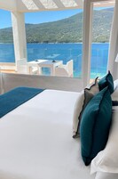 Quarto com cama, almofadas brancas e vista para o mar e montanhas.