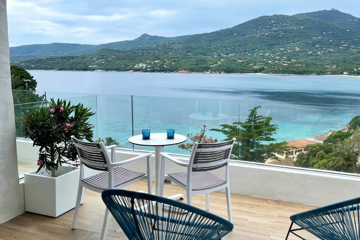 Un balcon avec une table et des chaises, donnant sur un paysage côtier avec une montagne en arrière-plan.