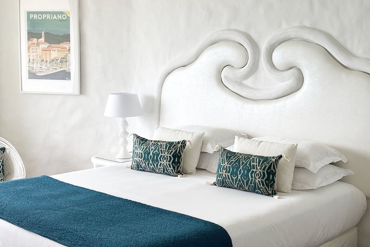 Un lit bien fait, avec du linge de lit blanc et deux coussins à motifs, un plaid turquoise et une tête de lit blanche ornée d’un motif sculpté.