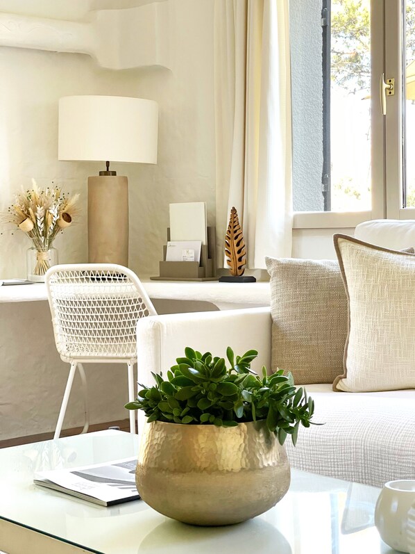 Un salón moderno con un sofá blanco, una mesa de centro de cristal, una silla blanca, una lámpara y un jarrón con plantas secas.