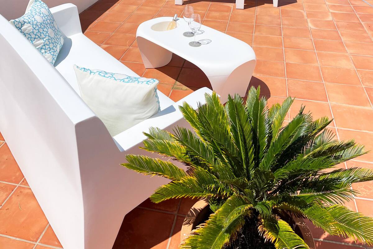 Une terrasse avec des meubles blancs, une table avec des boissons et une vue sur la mer et les montagnes.