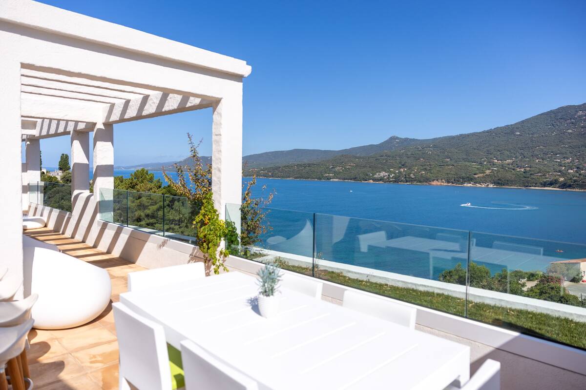 Une terrasse dotée de mobilier blanc et d’une balustrade en verre, offrant une vue sur une étendue d’eau et des collines.