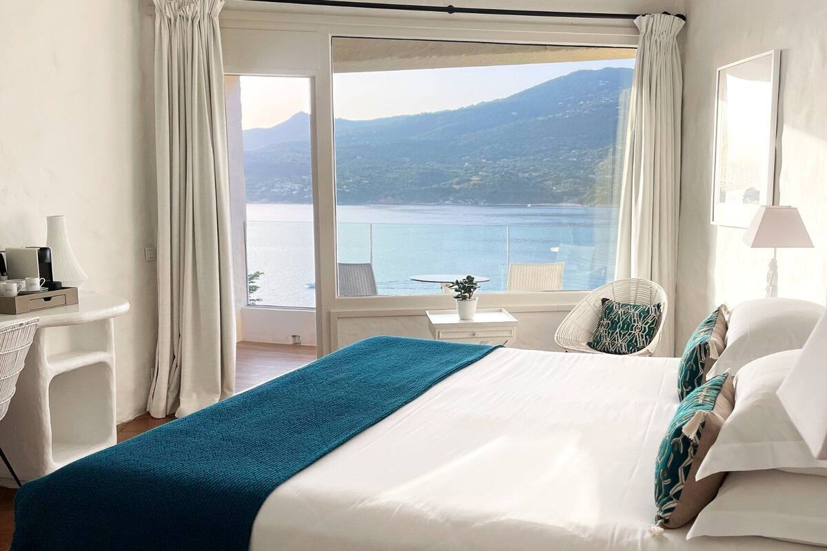 Une chambre d’hôtel avec un lit, une chaise, une petite table et une vue sur un plan d’eau et des montagnes.