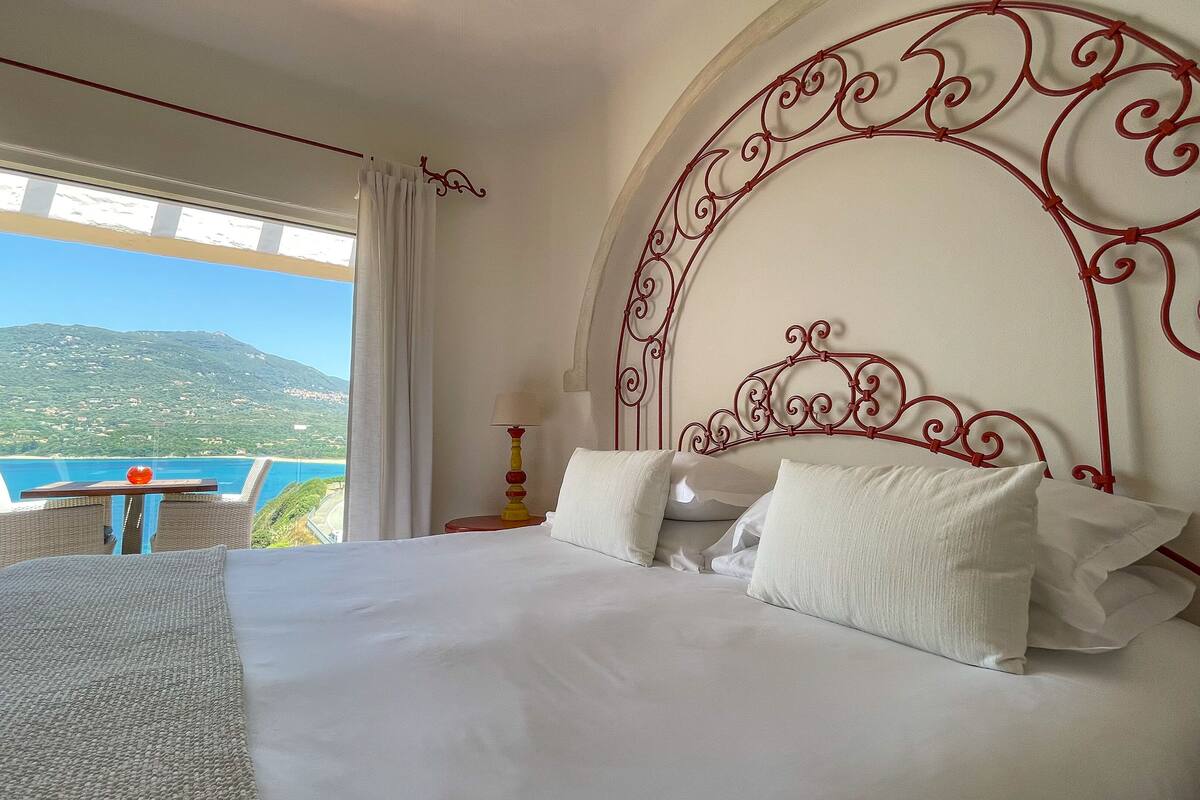 Une chambre d’hôtel avec un grand lit, une vue sur la montagne et la mer, et un balcon équipé d’une table et d’une chaise.