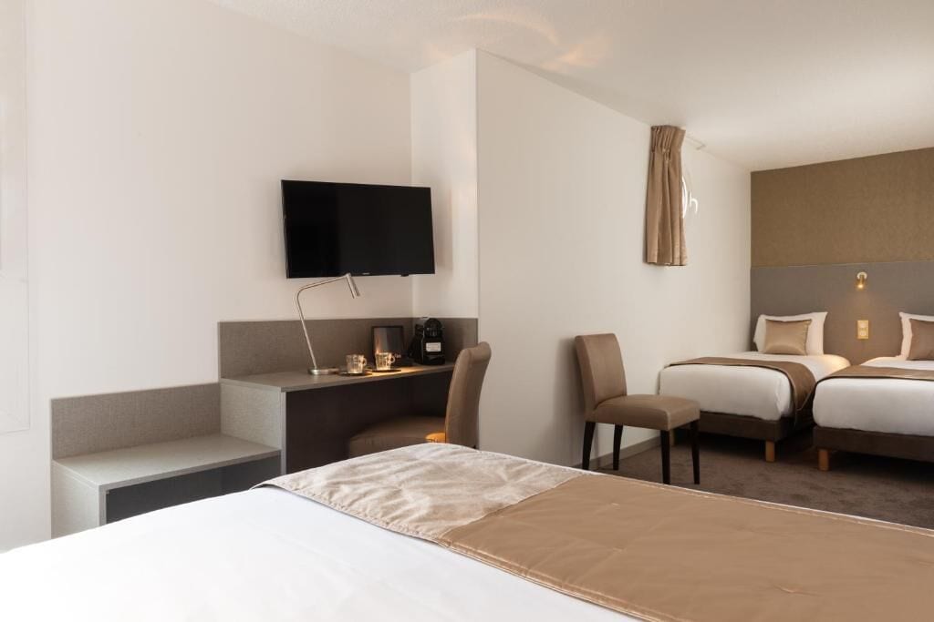 A hotel room with a bed, a desk with a chair, a television mounted on the wall, and two beds in the background.
