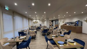 A modern dining area with multiple tables and chairs, a kitchen area with a coffee machine, and a large window with curtains. - The Originals City, Hôtel Orléans Nord (Saran)