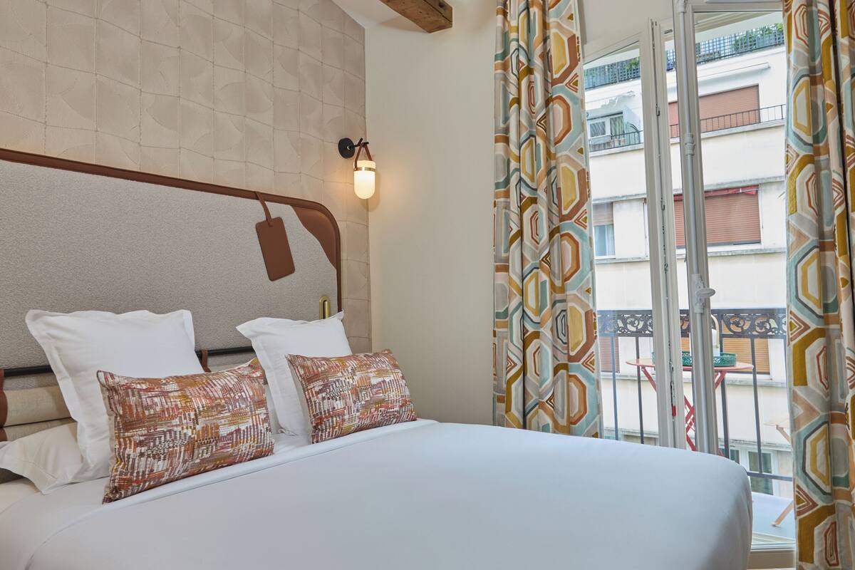 A hotel room with a large bed, a ceiling fan, a wall-mounted light, and a view of the outside balcony.