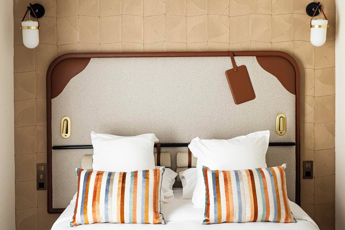 Quarto de hotel com uma cama grande, duas mesas de cabeceira, um painel de cabeceira com etiqueta e duas luminárias fixadas na parede.