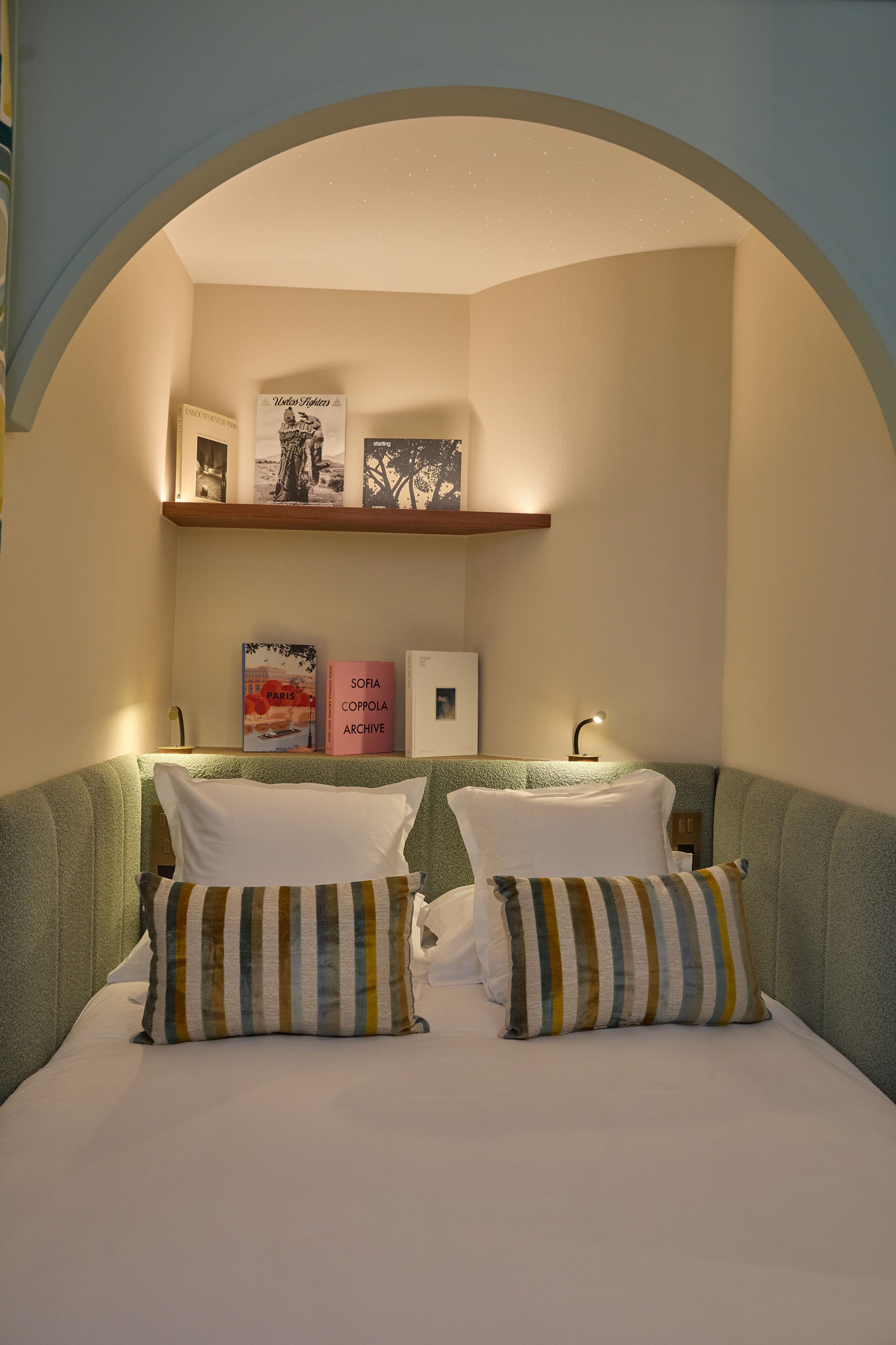 Un dormitorio con cama, almohadas, una mesita de noche, una estantería con libros y un cuadro colgado en la pared.