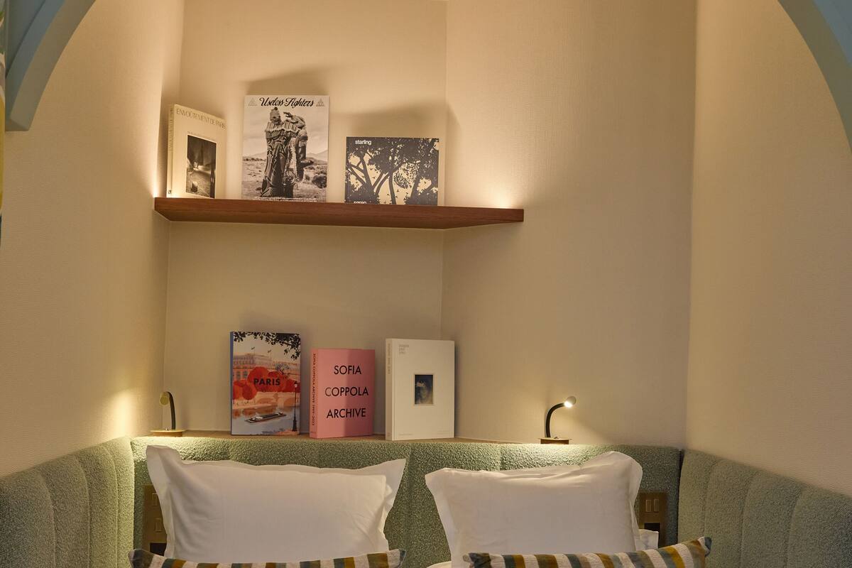 A bedroom with a bed, pillows, a nightstand, a shelf with books, and a wall-mounted picture.