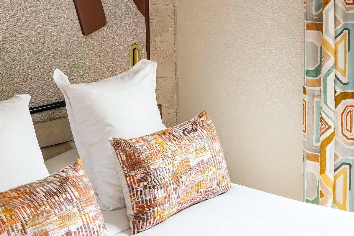A hotel room with a bed, pillows, a wall-mounted lamp, and a patterned curtain.