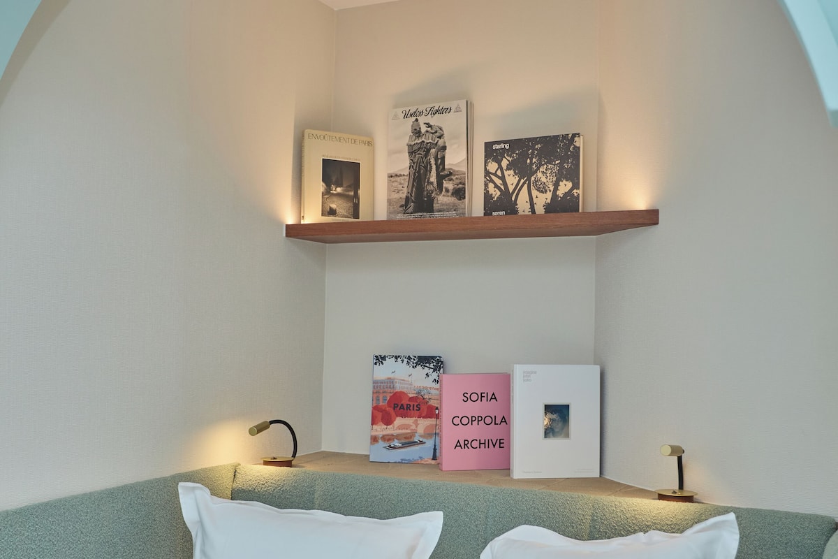 A bedroom with a bed, pillows, a shelf with books, and a wall arch.
