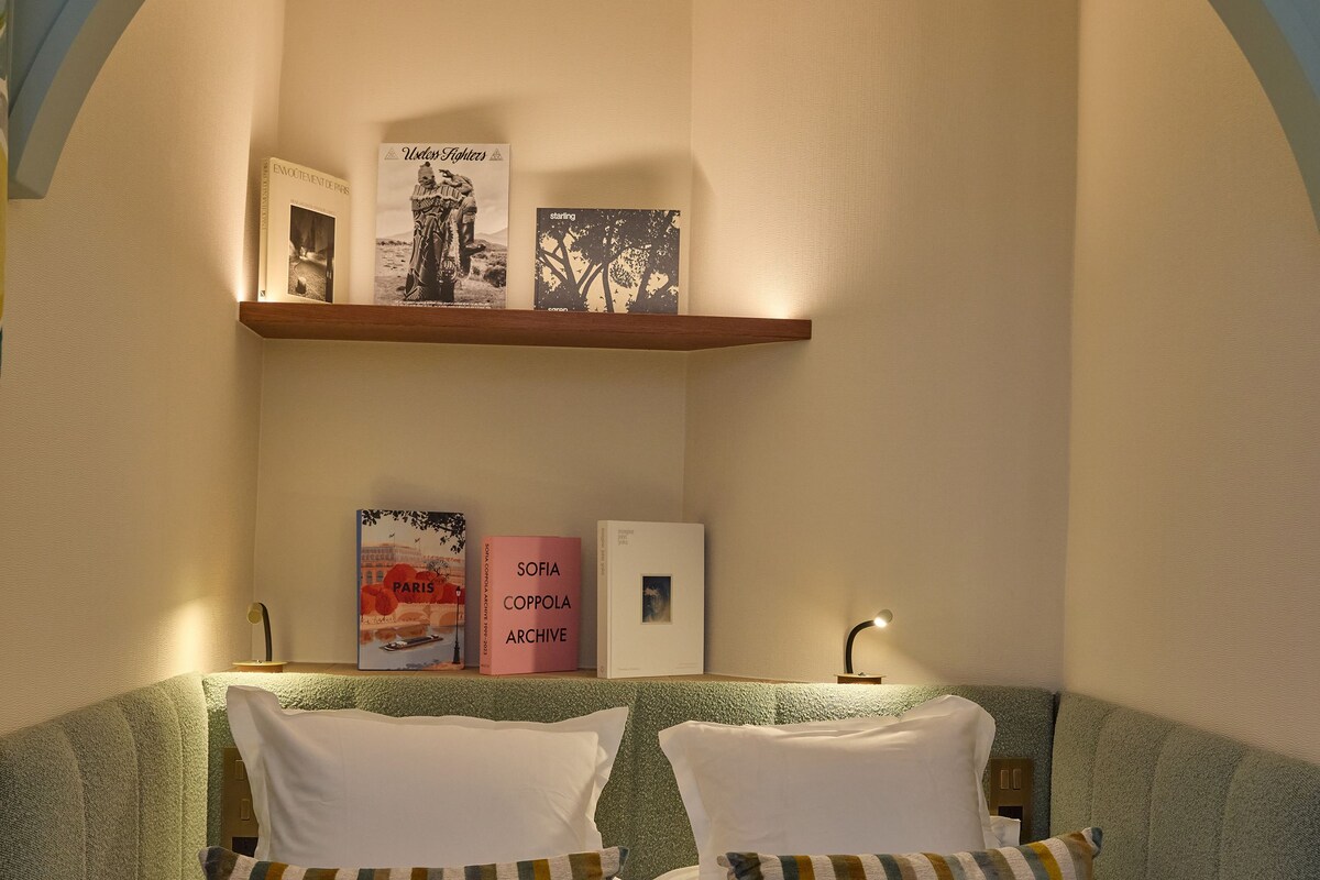 Un dormitorio con cama, almohadas, una mesita de noche, una estantería con libros y un cuadro colgado en la pared.