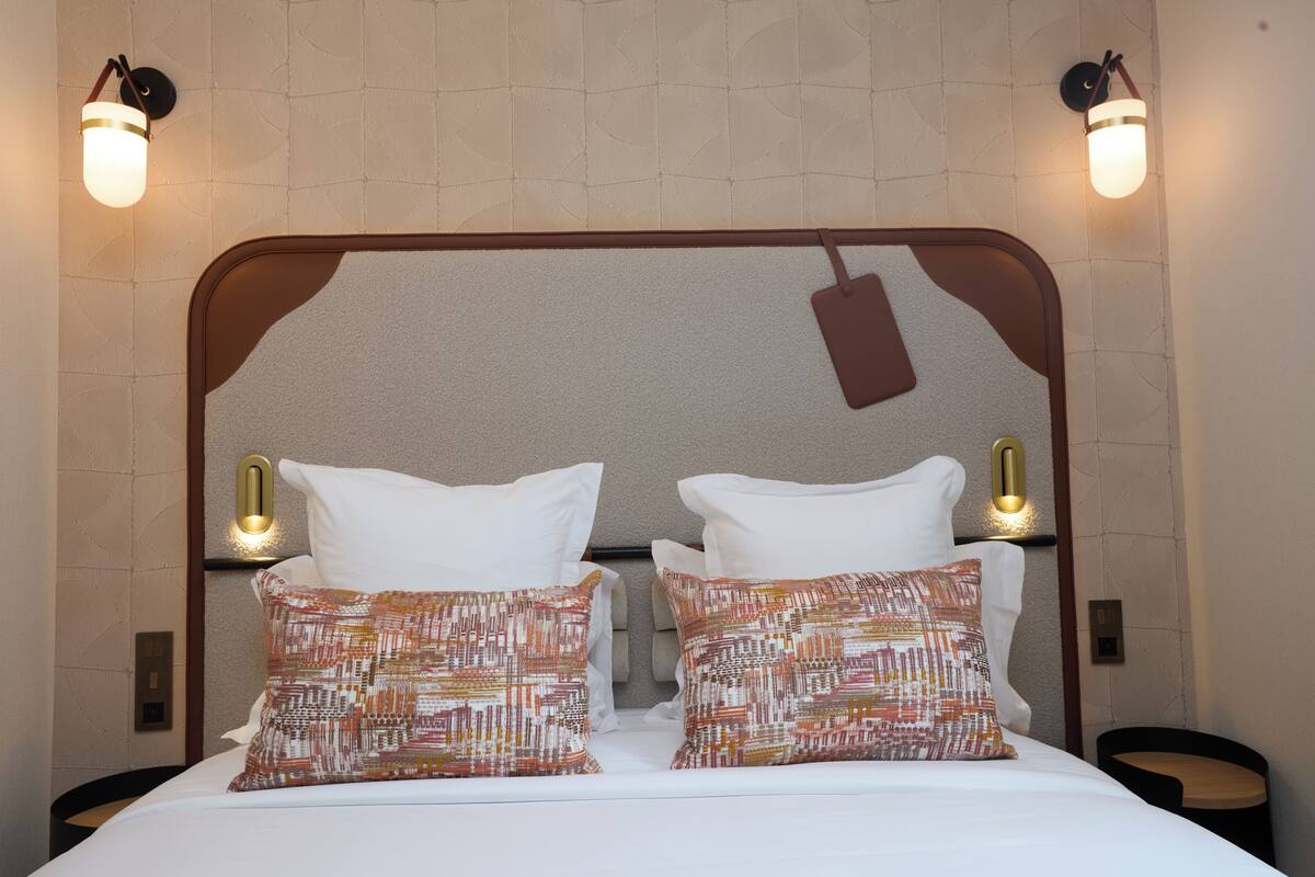 A hotel room with a large bed, two pillows, two wall-mounted light fixtures, and a small round table.
