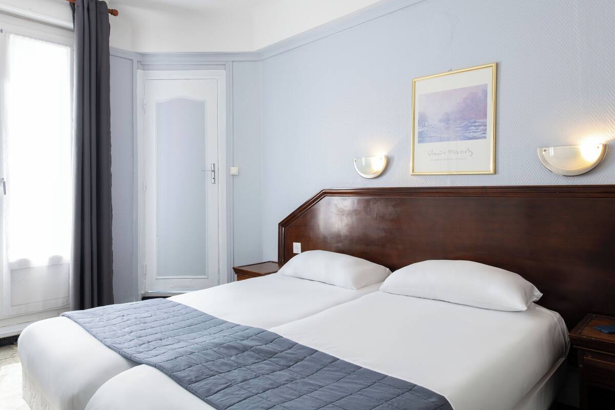 Hotelkamer met een tweepersoonsbed, nachtkastje, wandlampje, een lijst aan de muur en een raam met gordijnen.