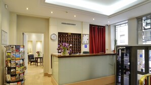 A hotel reception area with a reception desk, a clock, a bulletin board, and a magazine rack. - Hôtel Ambassadeur (Paris)