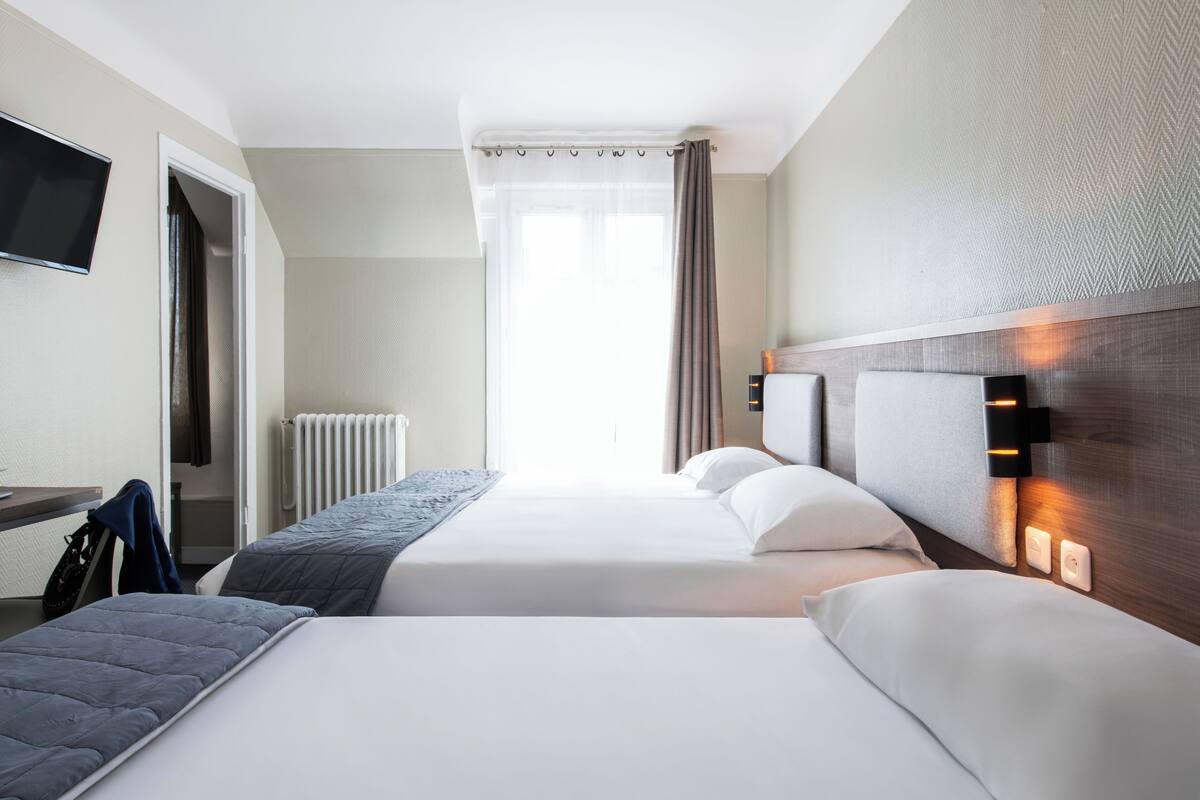 Een hotelkamer met twee bedden, een aan de muur bevestigde televisie, een raam met gordijnen en een radiator.