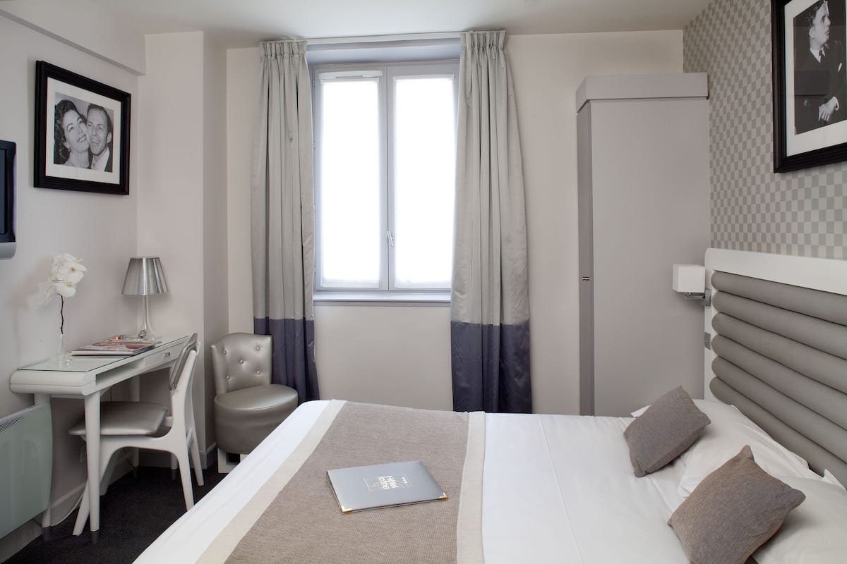 Quarto de hotel com cama, escrivaninha, cadeira e janela com cortinas.
