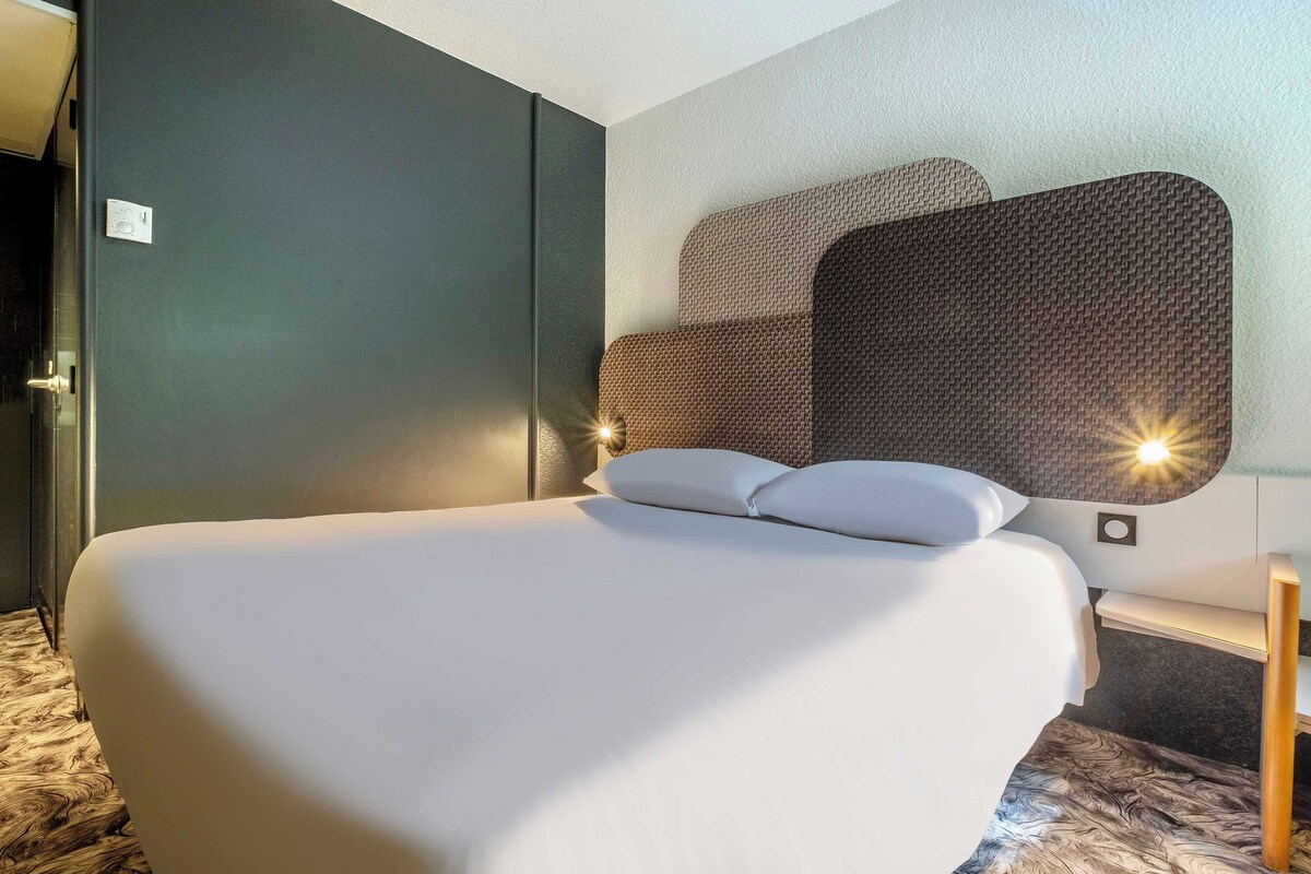 A hotel room with a large bed, a headboard with a textured design, and a bedside table with a lamp.