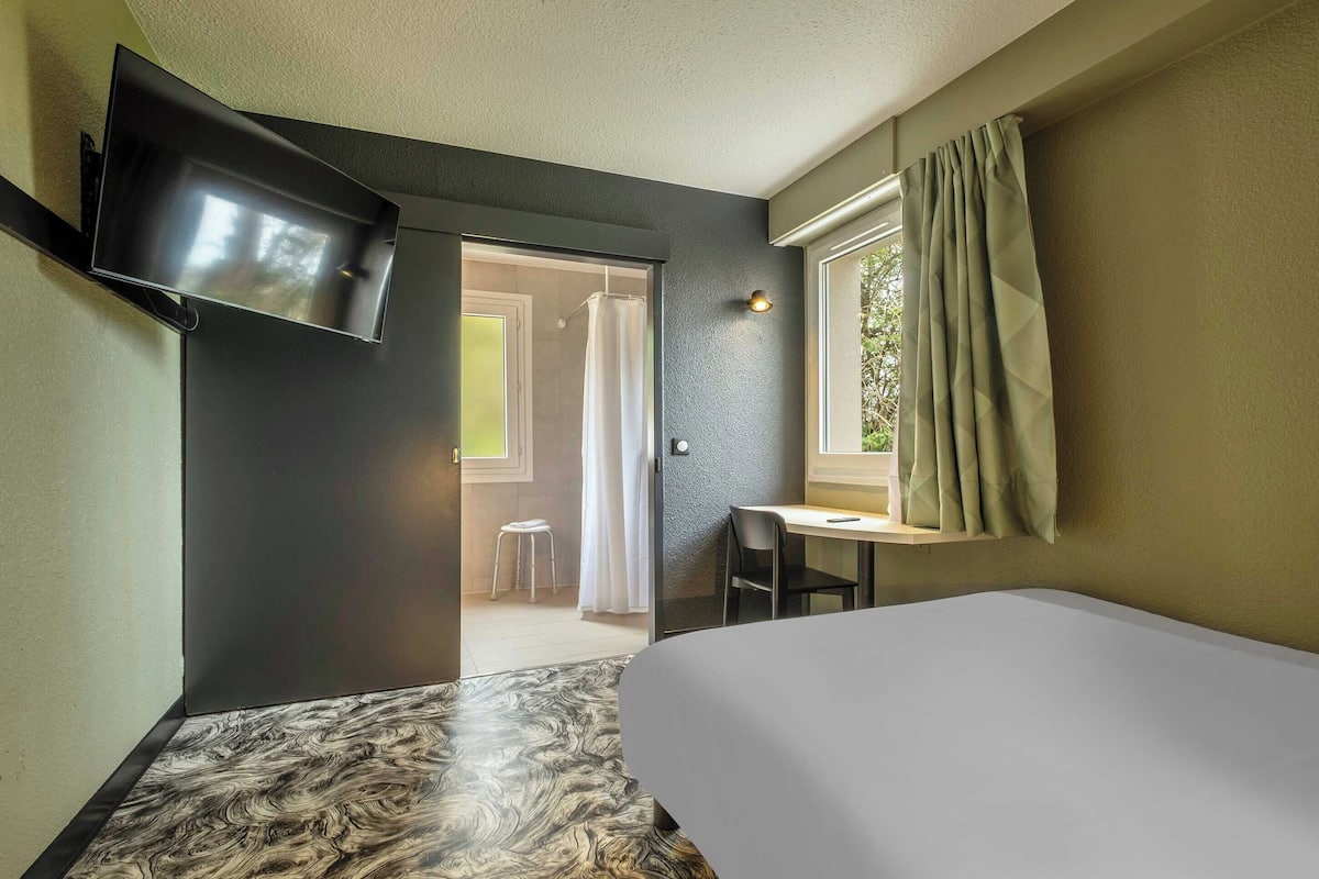 Ein Hotelzimmer mit Bett, Schreibtisch, Stuhl, Wand-Fernseher und einem Fenster mit Vorhängen.