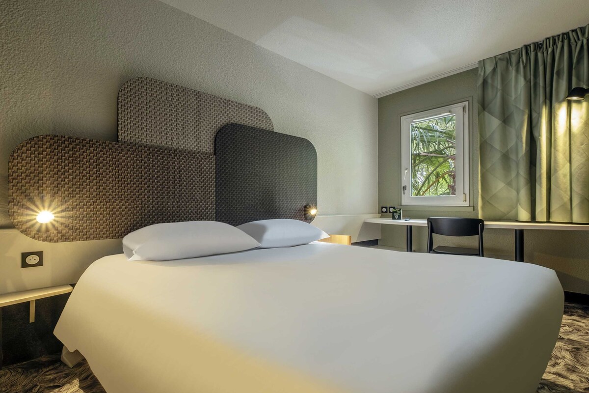 A hotel room with a large bed, a desk, a chair, and a window with curtains.