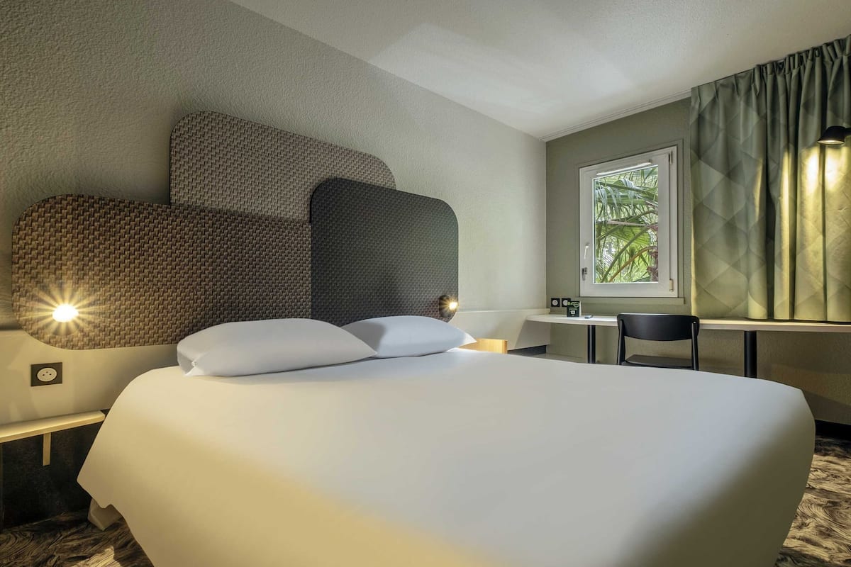 Ein Hotelzimmer mit einem großen Bett, einem Schreibtisch, einem Stuhl und einem fensterbepflanzten Fenster.