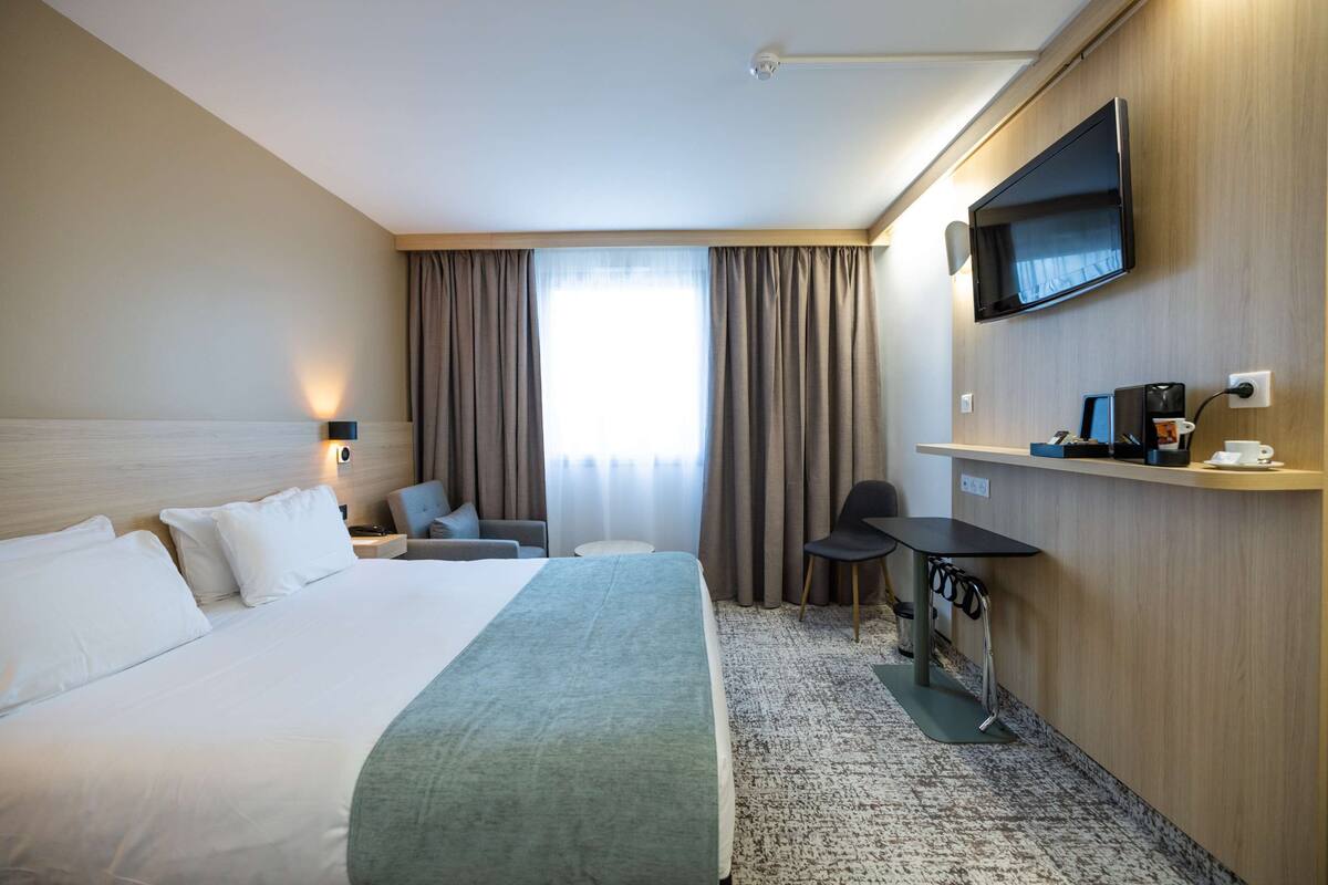 Una habitación de hotel con cama, escritorio, silla, televisión y ventana con cortinas.