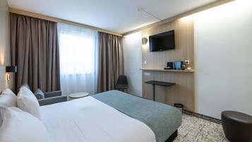 Une chambre d’hôtel avec un grand lit, un bureau, une chaise, un téléviseur fixé au mur et une fenêtre avec des rideaux.
