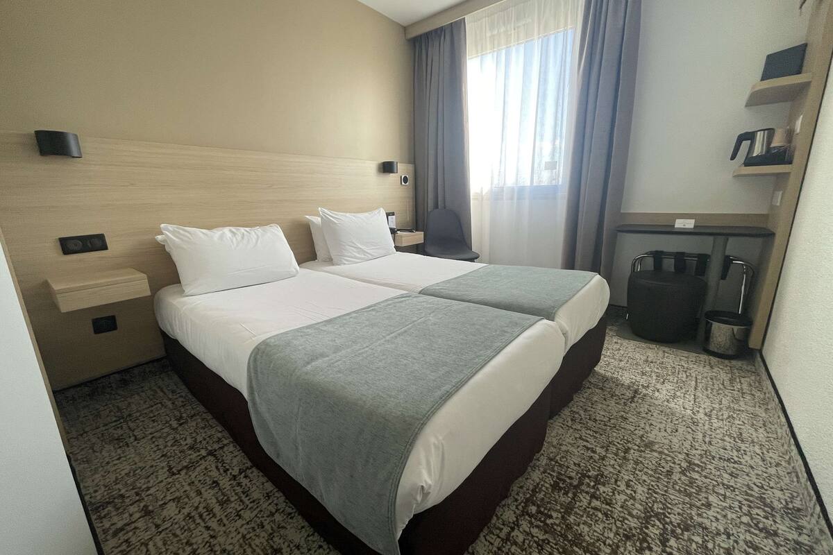 Habitación de hotel con una cama doble, un escritorio con silla, un cesto de basura pequeño y una ventana con cortinas.