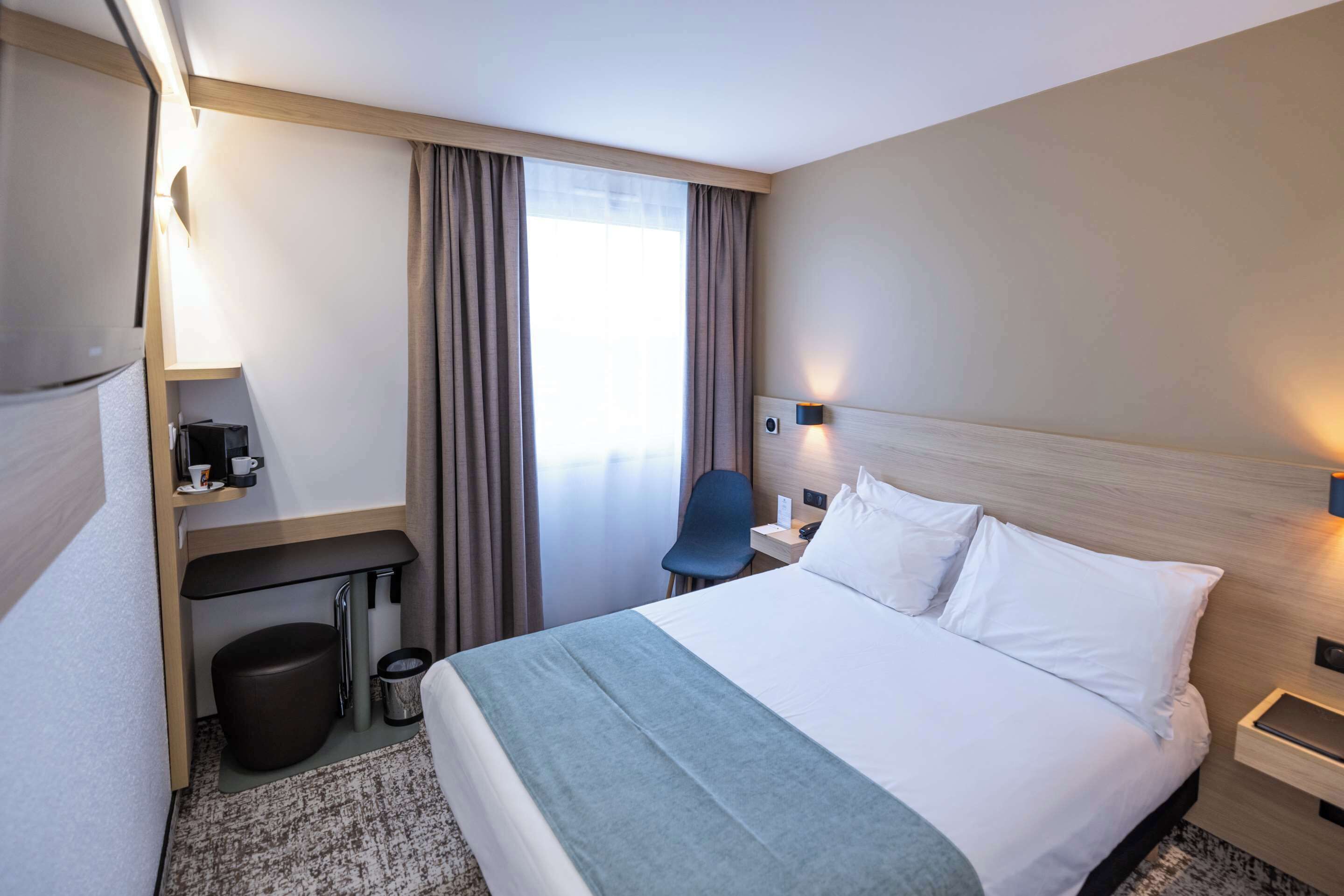 A hotel room with a bed, a desk, a chair, a trash bin, and a window with curtains.