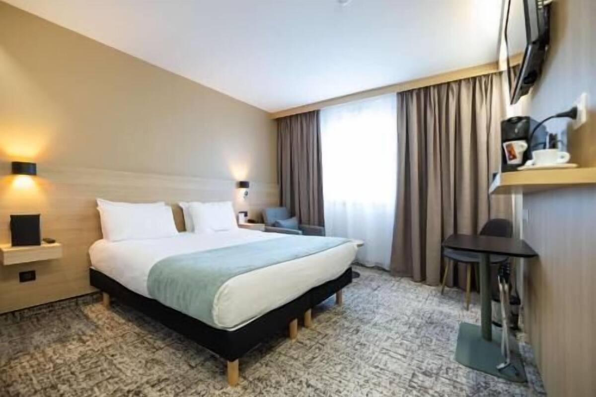 Habitación de hotel con cama, mesitas de noche, escritorio, silla, televisión y ventana con cortinas.
