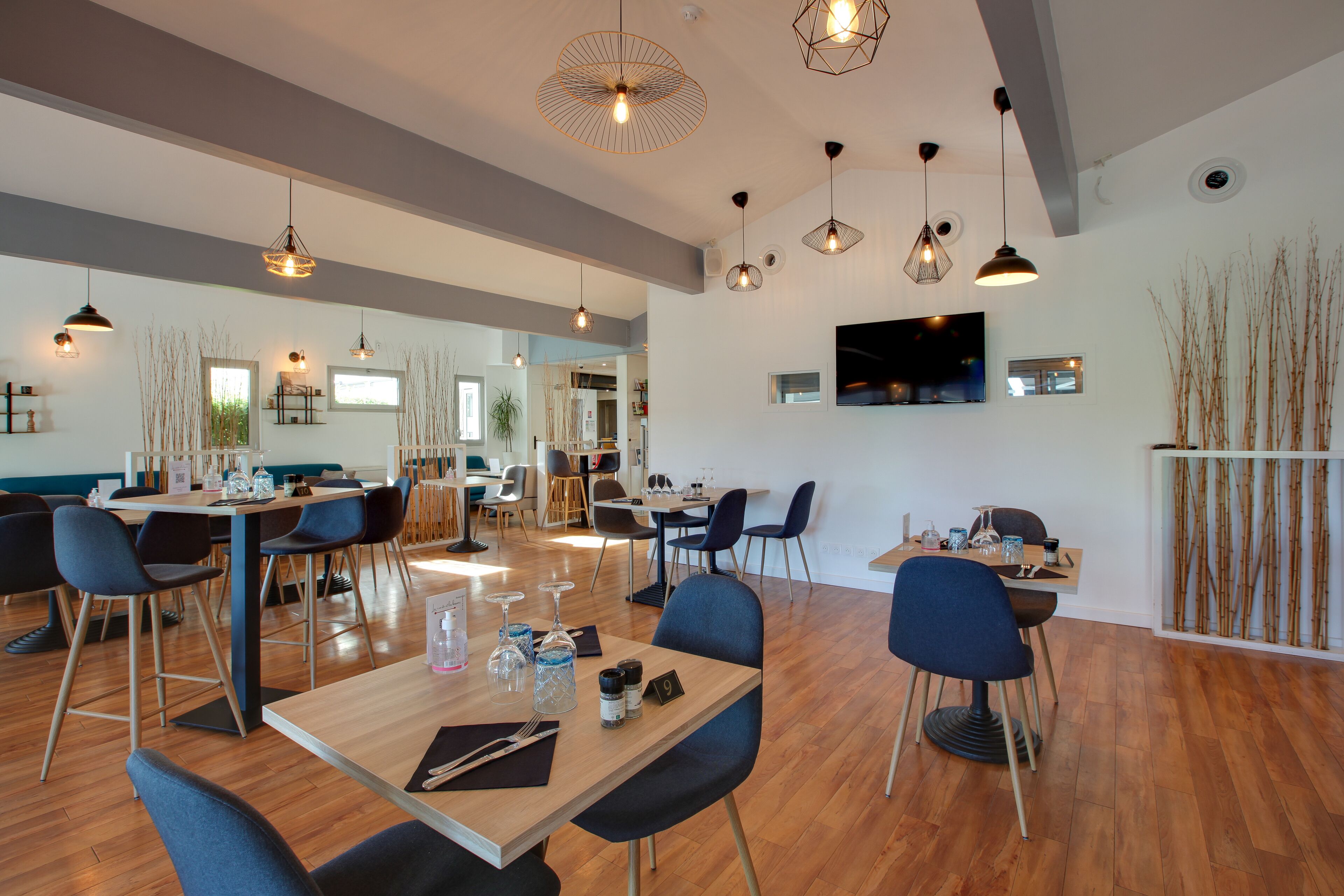 A modern dining area with wooden tables and blue chairs, a flat-screen TV mounted on the wall, and a large window providing natural light.