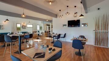 A modern dining area with wooden tables and blue chairs, a flat-screen TV mounted on the wall, and a large window providing natural light.