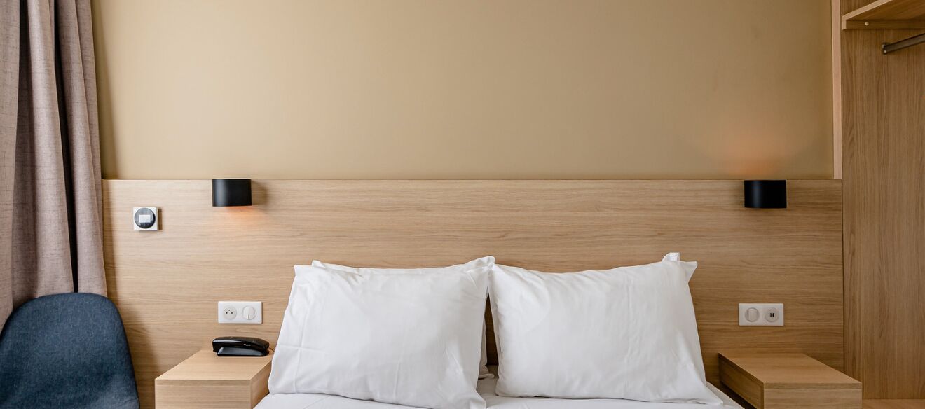 Hotelkamer met houten hoofdeinde, twee witte kussens, nachtkastje met telefoon en een wandlamp.