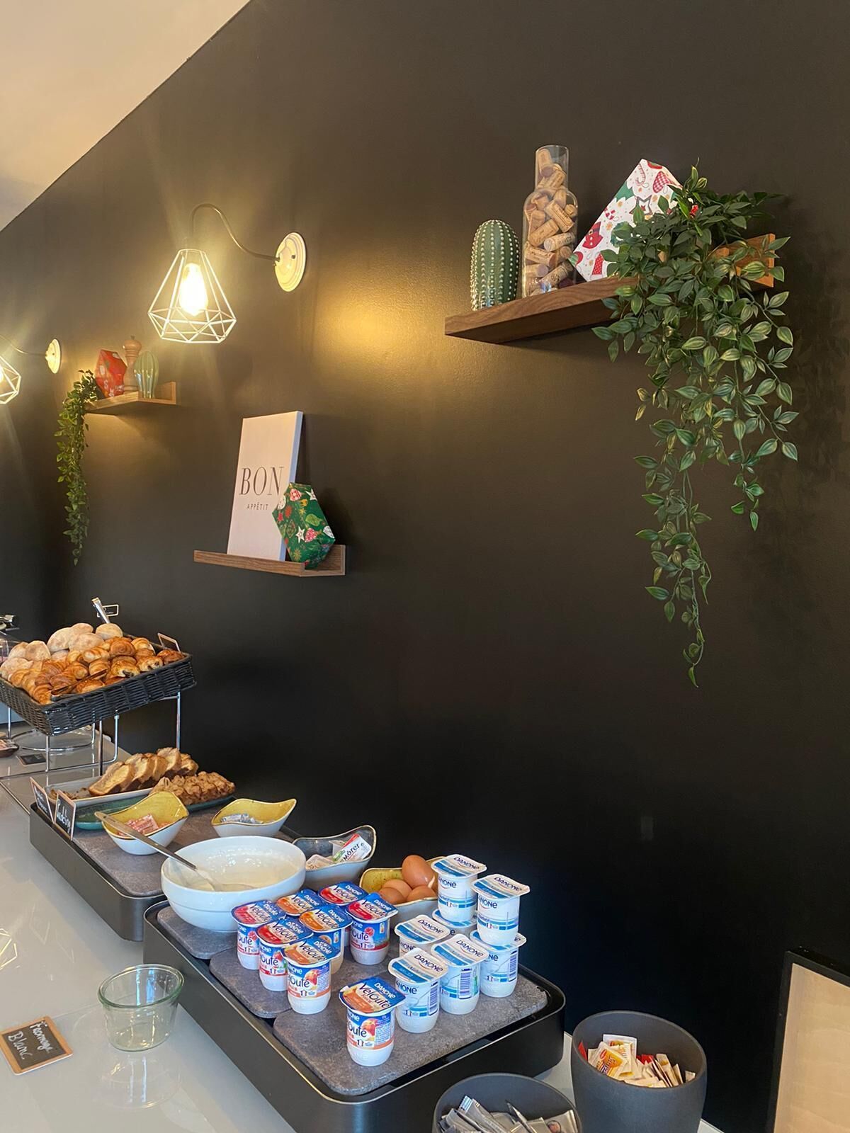 A breakfast buffet with various food items, including pastries, bread, and dairy products, arranged on a counter with decorative lighting and plants.