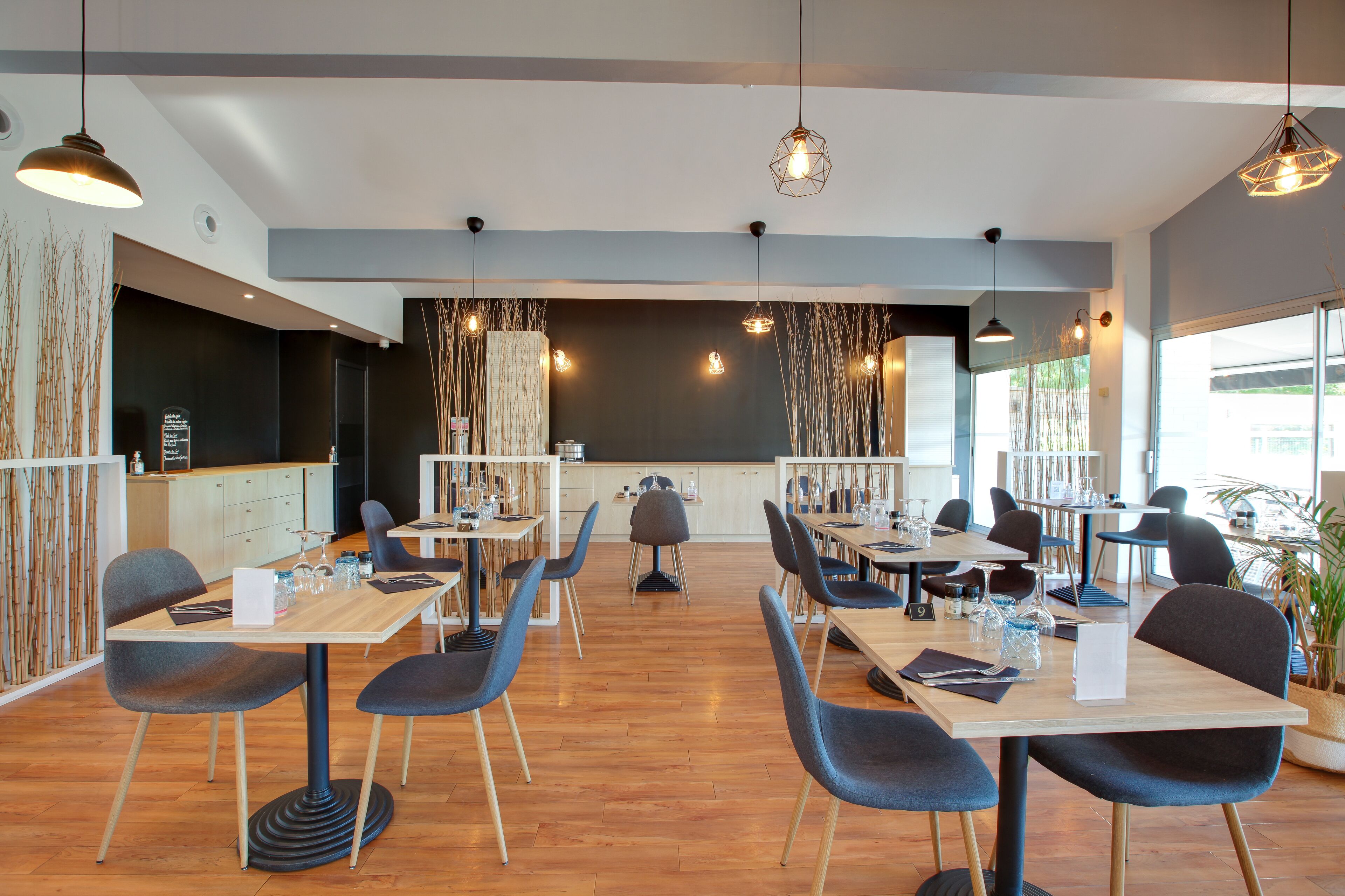 Een modern restaurant met houten tafels en stoelen, hangende verlichting en een menubord op een schoolbord.