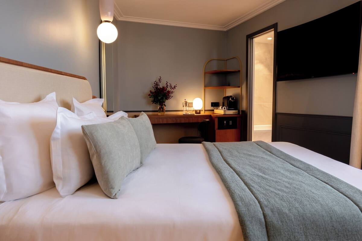 A hotel room with a neatly made bed, a desk with a lamp, a coffee maker, and a TV on the wall.